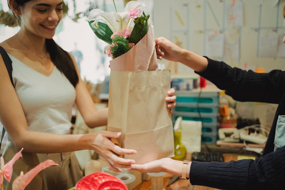 A florist woman with long dark hair, wearing a white sleeveless top, is smiling as she receives a floral bouquet wrapped in pink paper, arranged with white lilies, pink roses, and greenery, from anoth
