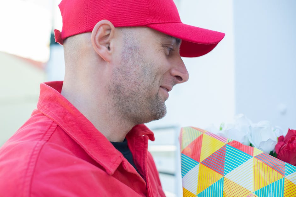 A delivery person wearing a red cap and red jacket is shown in profile, holding a floral gift wrapped in vibrant, geometric-patterned paper with yellow, blue, pink, and white triangles, and a white ri