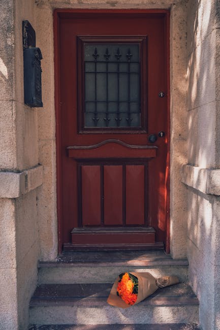 A bouquet of fresh flowers, including vibrant orange roses, yellow chrysanthemums, and lush greenery, wrapped in brown kraft paper and secured with a natural twine ribbon, rests on the stone doorstep 