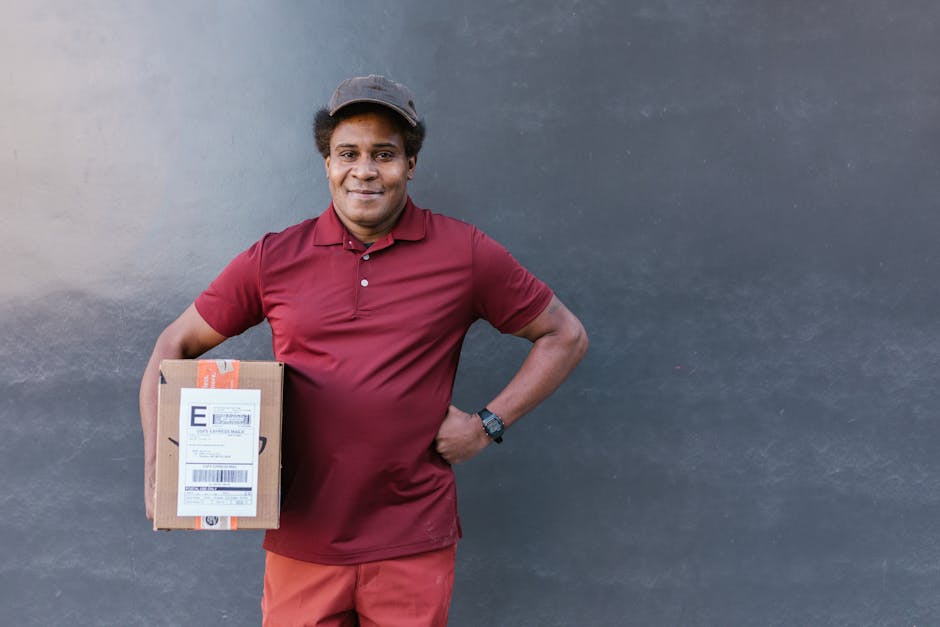 A smiling male flower delivery courier with dark skin, wearing a maroon polo shirt, matching maroon trousers, and a grey cap, stands outdoors against a plain, dark grey wall. He holds a medium-sized c