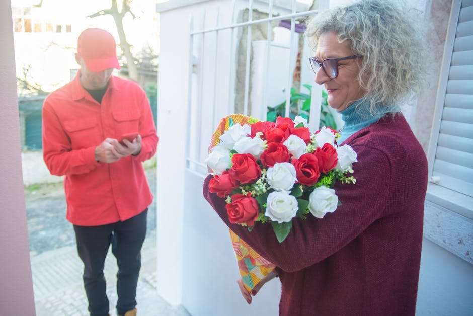 A woman with curly gray hair and glasses is holding a vibrant floral bouquet featuring red and white roses wrapped in patterned paper, standing near a doorway with natural daylight illuminating her fa