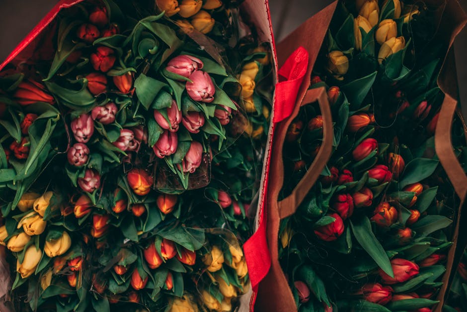 A vibrant display of fresh tulip bouquets featuring pink, yellow, and red flowers, neatly arranged in large brown paper and fabric shopping bags. The tulips have lush green stems and leaves, with thei