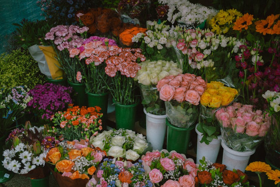 A vibrant display of fresh floral arrangements and bouquets at a flower shop, featuring roses in soft pink, yellow, and peach, along with clusters of white, purple, and pink blossoms. The flowers are 