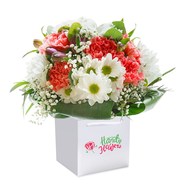 This superbly arranged bouquet, displayed upright in a clean, square white gift bag, beautifully embodies the cheerful spirit of Nunhead. Soft coral-pink and peach carnations fill the centre, their ruffled edges layered for sumptuous texture, while occasional bright red carnations add depth and visual excitement. Clusters of white daisy-like chrysanthemums, each with a striking lime-green centre, are thoughtfully interspersed for brightness-echoing the open, welcoming ambience of Nunhead's green spaces. Sprigs of delicate white baby's breath and hints of unopened purple-tipped buds provide refined touches, weaving softness and subtle intrigue throughout. The display is completed with luxurious, broad green foliage fanning and curving gracefully around the base, creating a lush 'collar' effect reminiscent of a walk through Nunhead's tree-shaded lanes. Designed with careful attention, the bouquet has a rounded silhouette, conveying abundance and balance-a hallmark of the area's trusted florists. The white gift bag, fitted with sturdy ribbon handles, is both modern and practical, perfect for surprise doorstep delivery or as a gleaming centrepiece at a Lancastre Road gathering. The entire composition is photographed against a pure white background, highlighting every vibrant hue and detail for maximum impact. Every element, from the gentle interplay of colours to the sleek branding and presentation, speaks of freshness, quality, and heartfelt craftsmanship. Ideal as a celebratory gift-be it for a birthday, thoughtful thank you, or a Nunhead anniversary-this arrangement encapsulates the beauty of local floristry, ready to bring joy to any occasion within this friendly south London neighbourhood.
